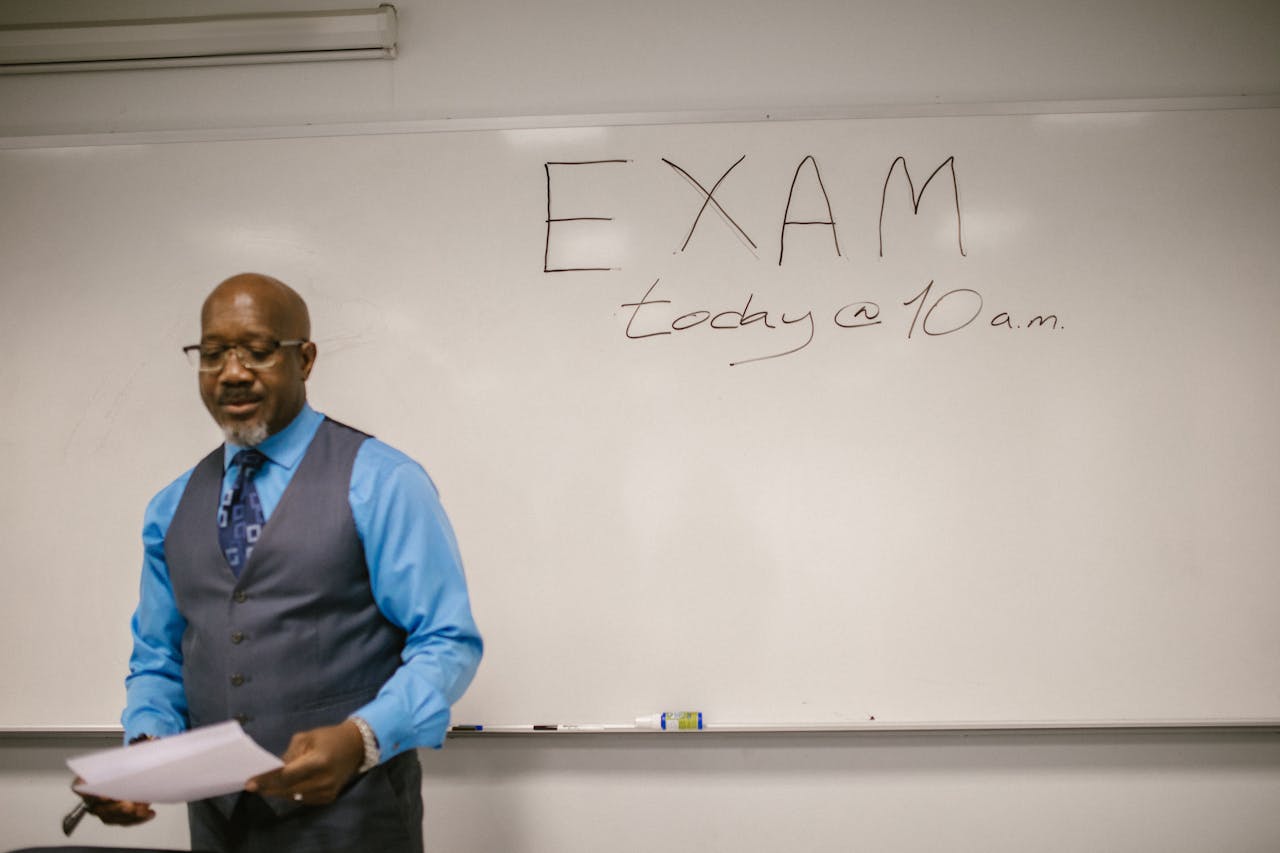 impact-img A professor stands next to a whiteboard with 'Exam today at 10 a.m.' written, indicating an upcoming exam in a classroom.
