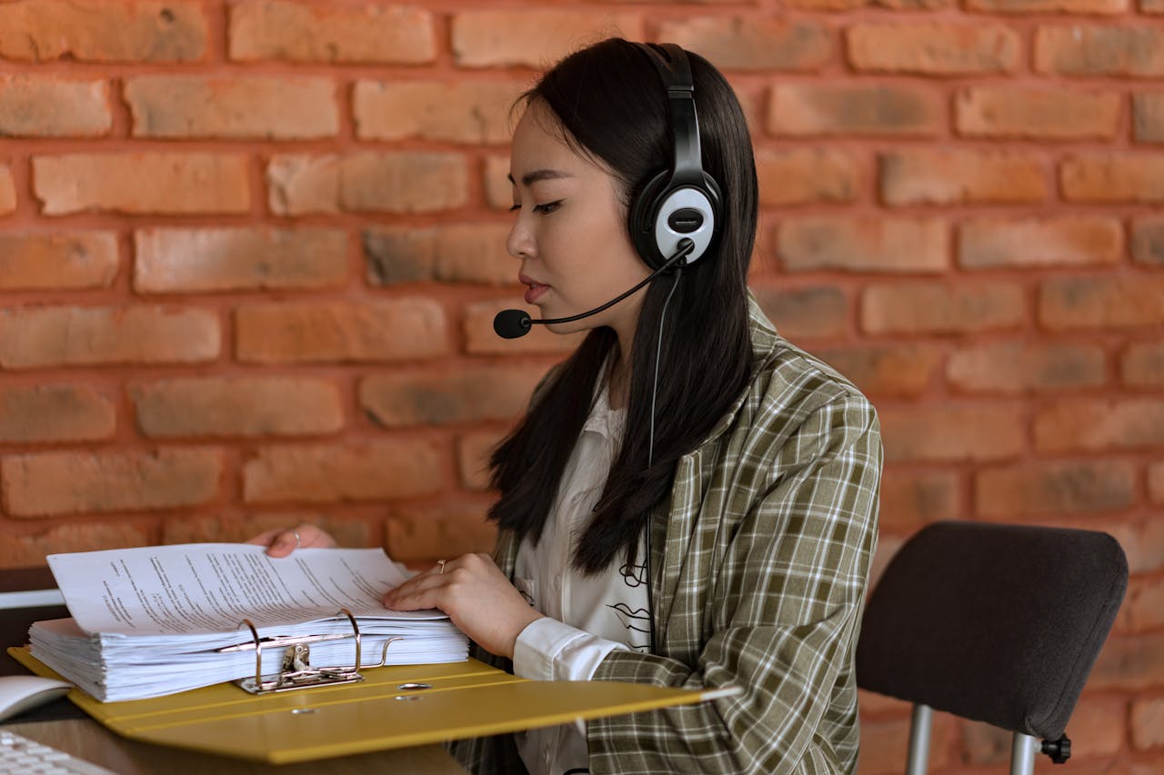 An Asian woman working in an office, wearing headphones and reviewing documents.